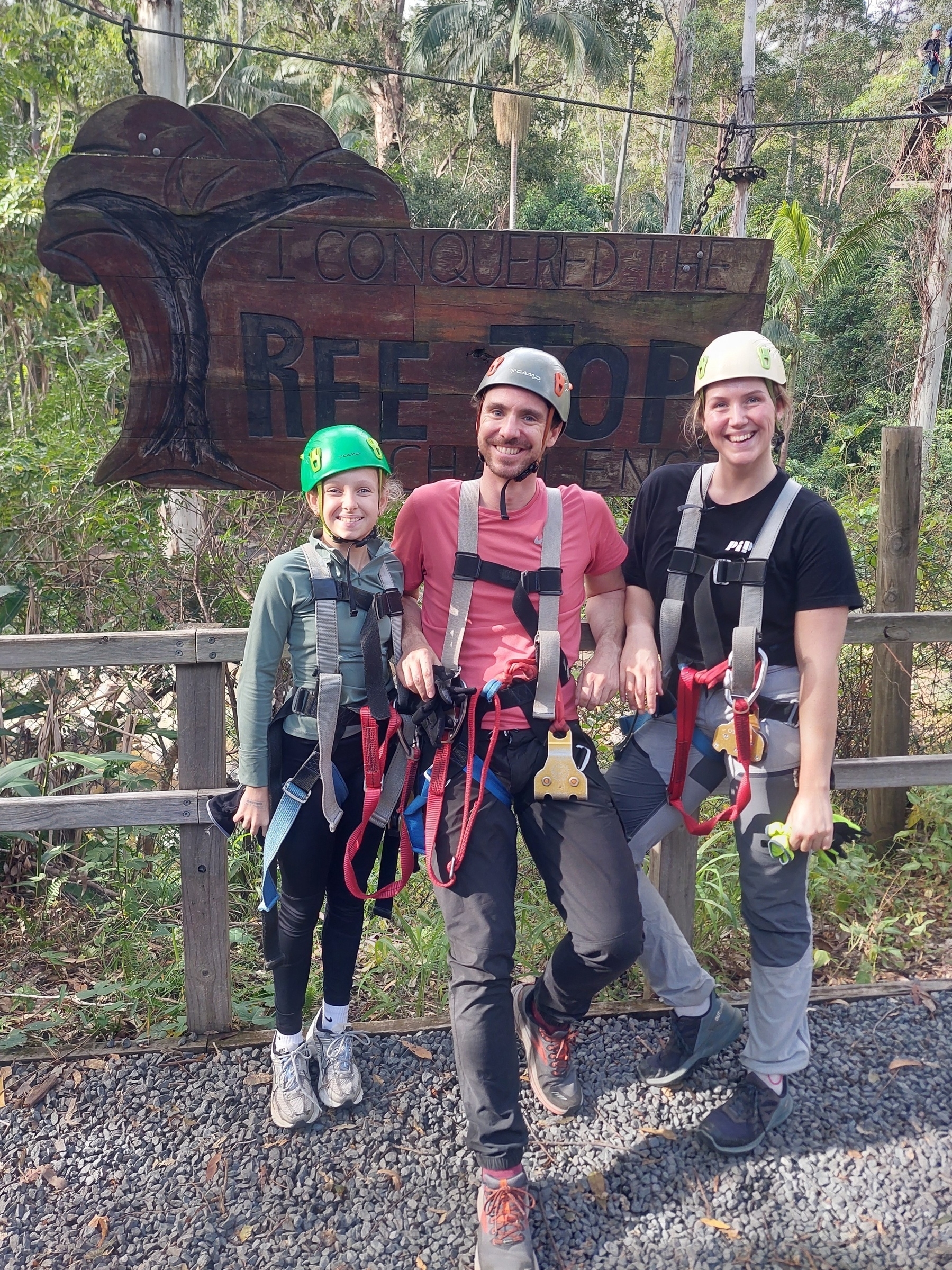 Auto-generated description: Tanja, Chanel and Joe in harnesses and helmets are smiling in front of a wooden sign in a forested area that reads, I conquered the treetops.