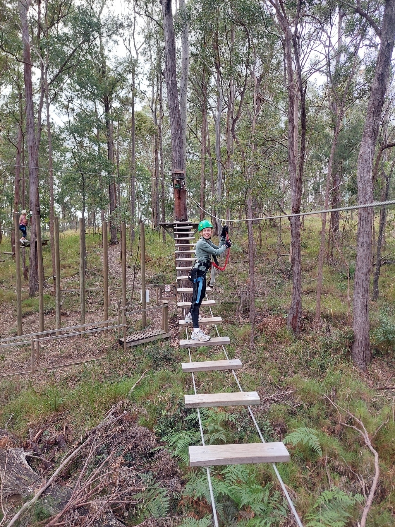 Auto-generated description: Chanel wearing a green helmet and harness navigates a rope bridge in a wooded adventure course.