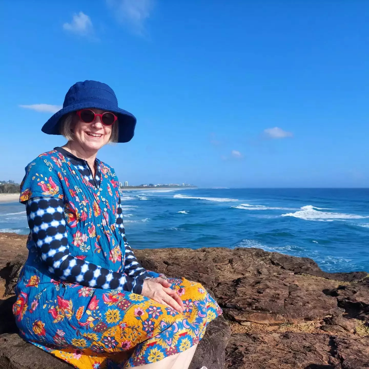Auto-generated description: A person wearing a colorful outfit and a blue hat is smiling while sitting on a rocky coastline with the ocean in the background.