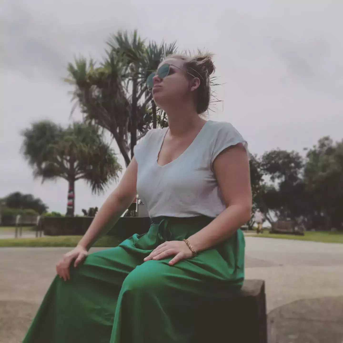 Auto-generated description: A person wearing sunglasses sits outdoors on a bench, dressed in a white shirt and green skirt, with palm trees in the background.