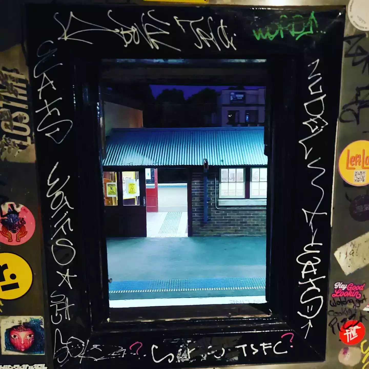 Auto-generated description: A window frame is covered in graffiti and stickers, overlooking a view of a small building with a corrugated metal roof.