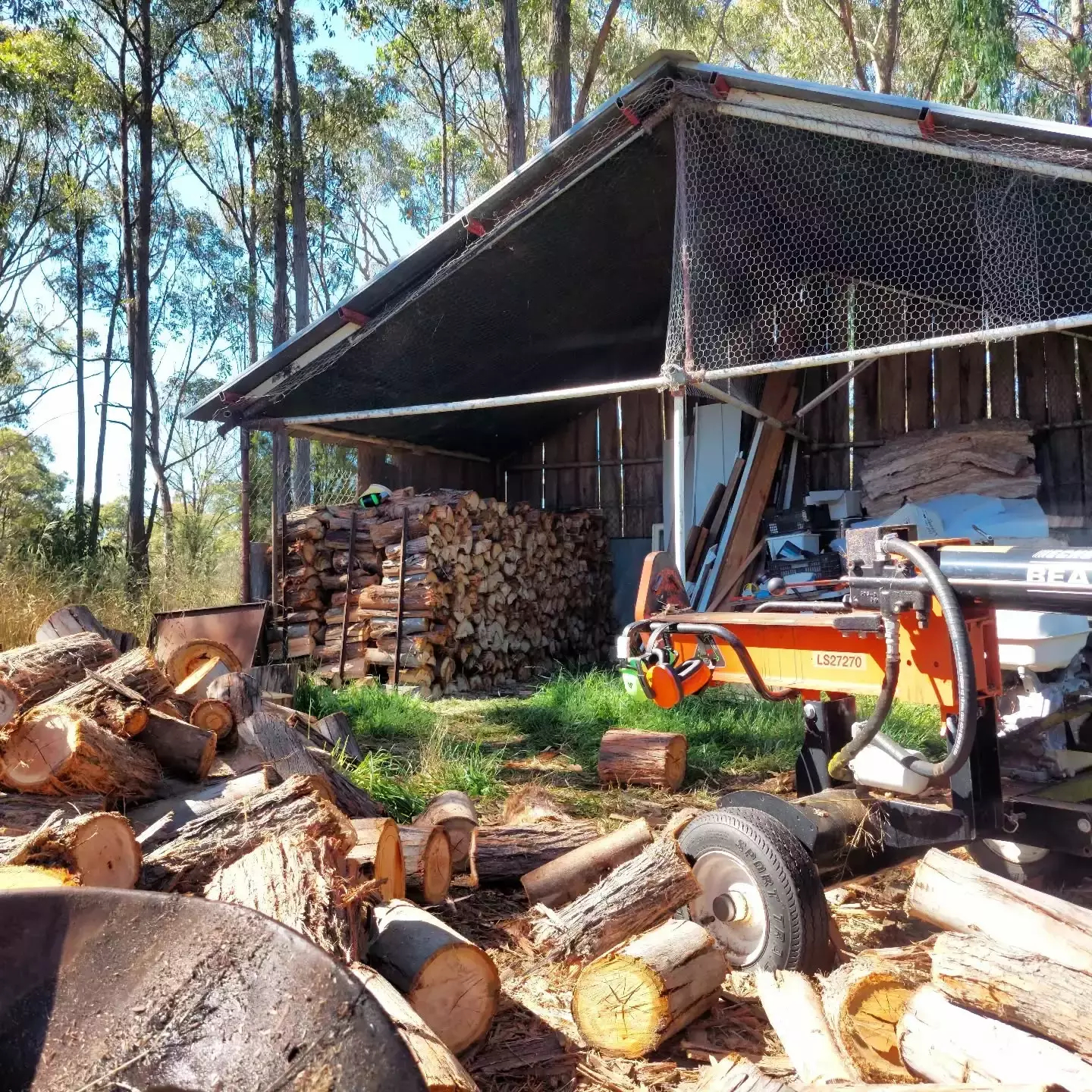 Auto-generated description: A log splitter is parked beside a stack of firewood under a rustic shelter, surrounded by chopped logs in a woodland area.