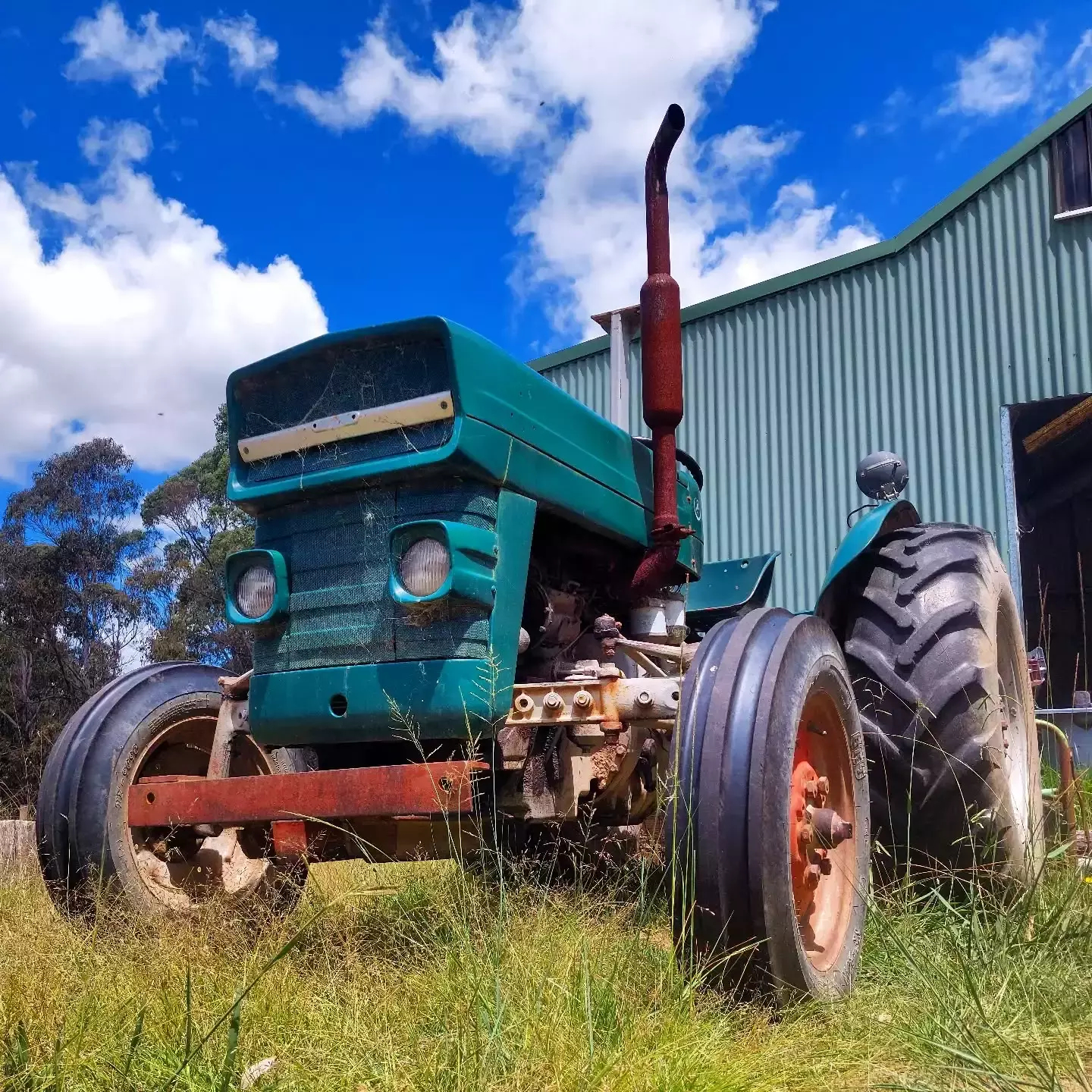 Auto-generated description: A green tractor is parked on grass in front of a corrugated metal building under a blue sky.