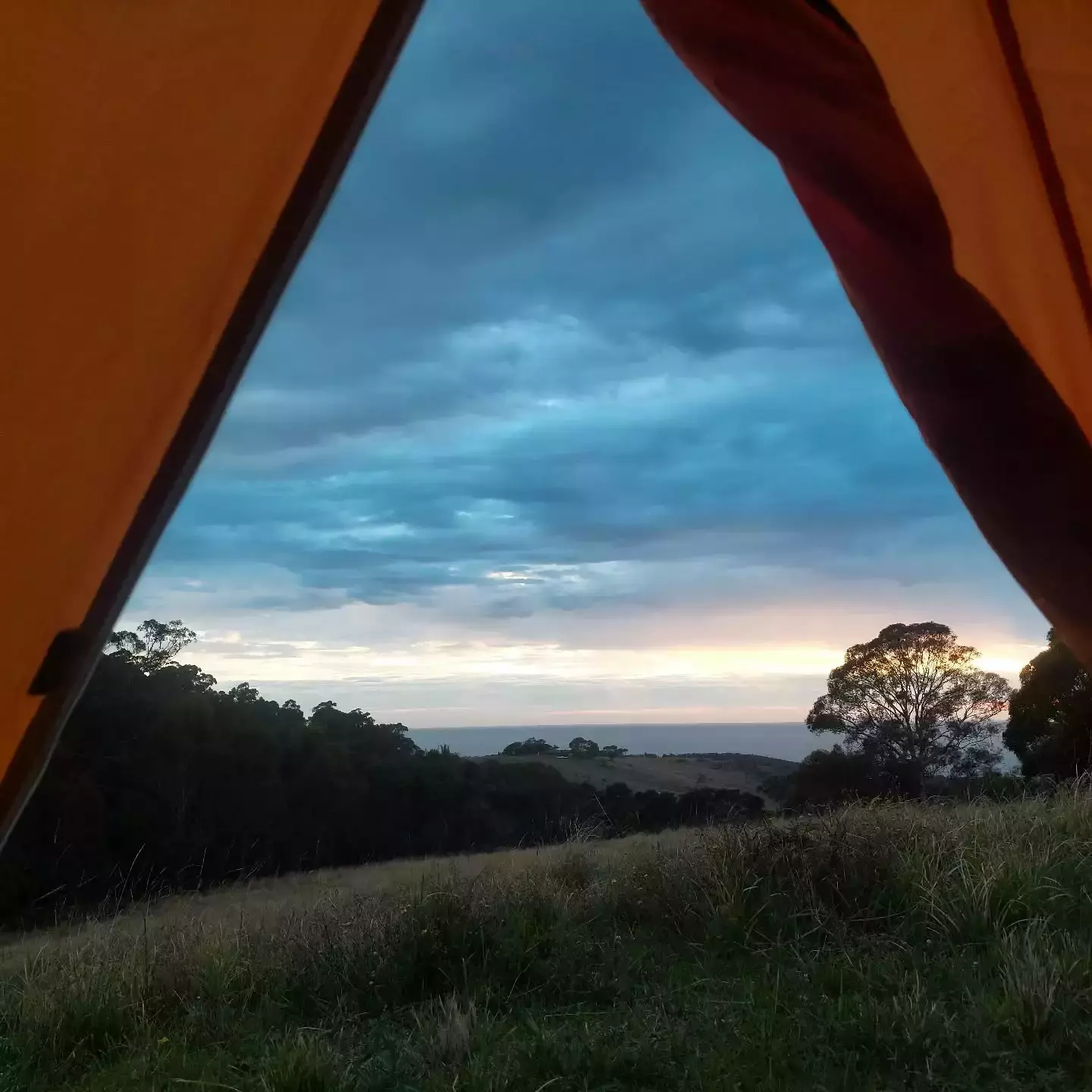 Auto-generated description: A view of the horizon with a vibrant sunset and rolling hills is seen from the opening of an orange tent.