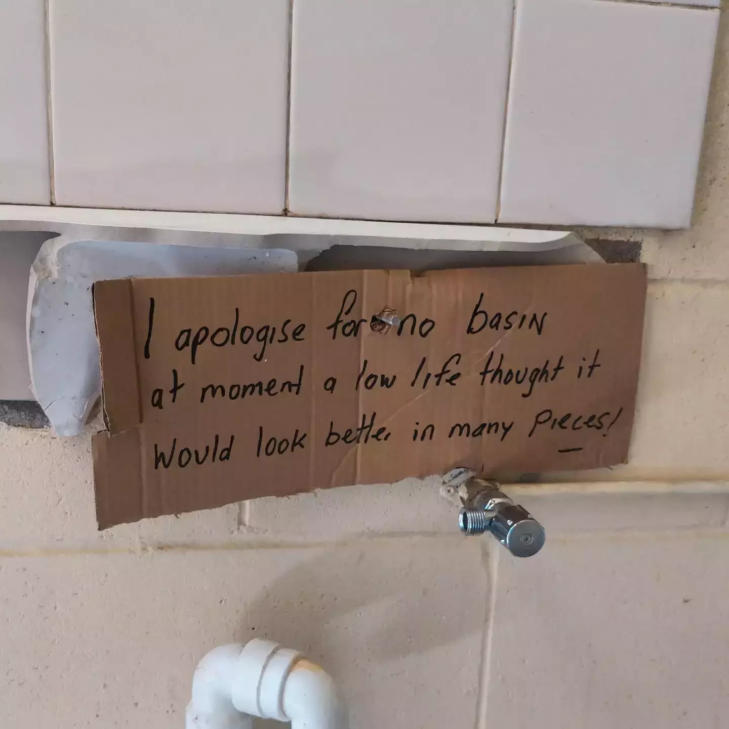 Auto-generated description: A handwritten sign is taped above plumbing pipes, apologizing for the absence of a basin due to someone breaking it.