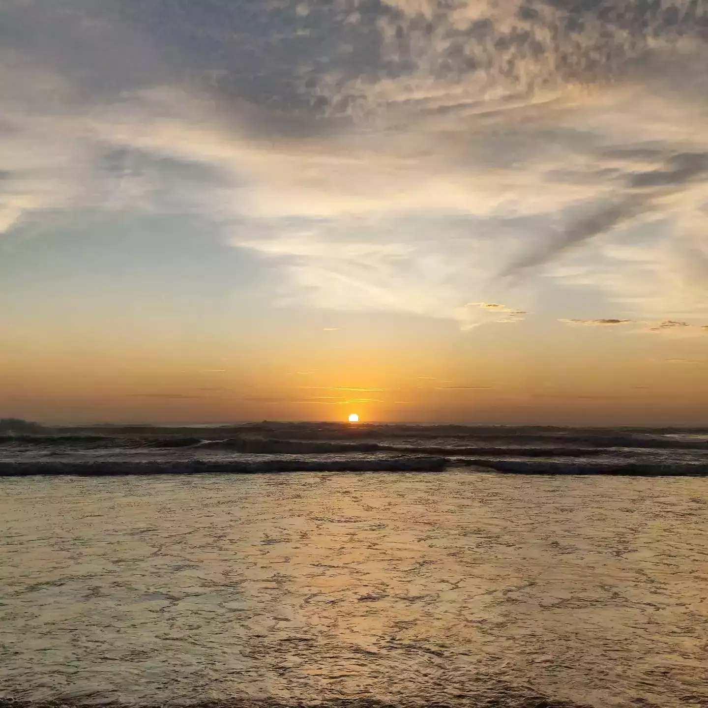 Auto-generated description: A serene sunset over the ocean with gentle waves and a cloudy sky.