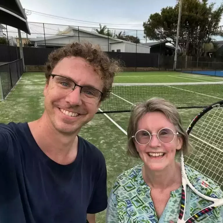 Auto-generated description: A man and a woman with tennis rackets pose for a selfie on a tennis court.