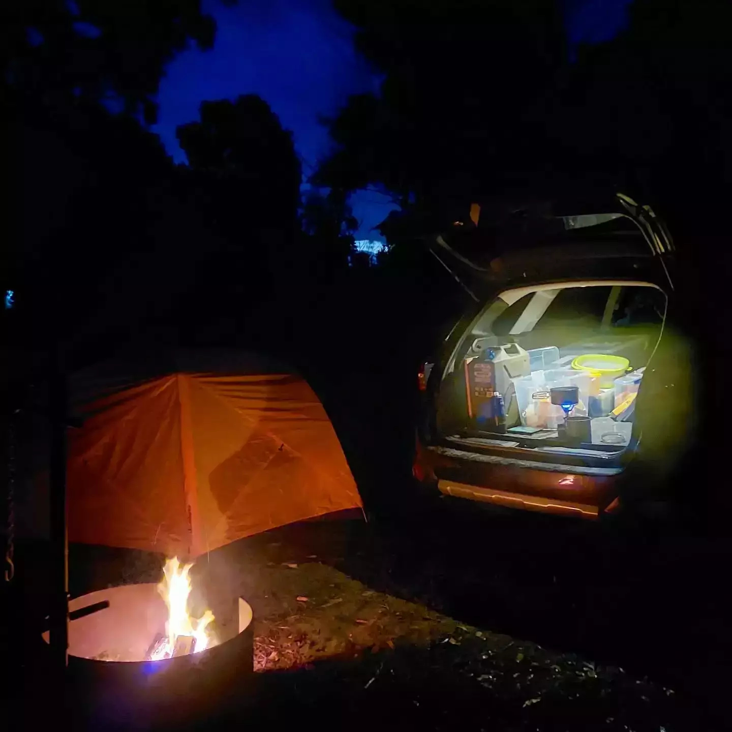 Auto-generated description: A nighttime camping scene with a lit campfire, an orange tent, and the open trunk of a car filled with camping gear.