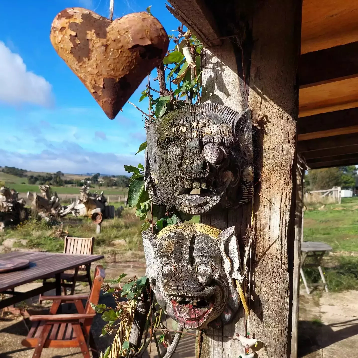 Auto-generated description: A rustic outdoor scene features two carved wooden masks on a post beside a hanging heart decoration, with a view of a wooden table and chairs set against a pastoral landscape.