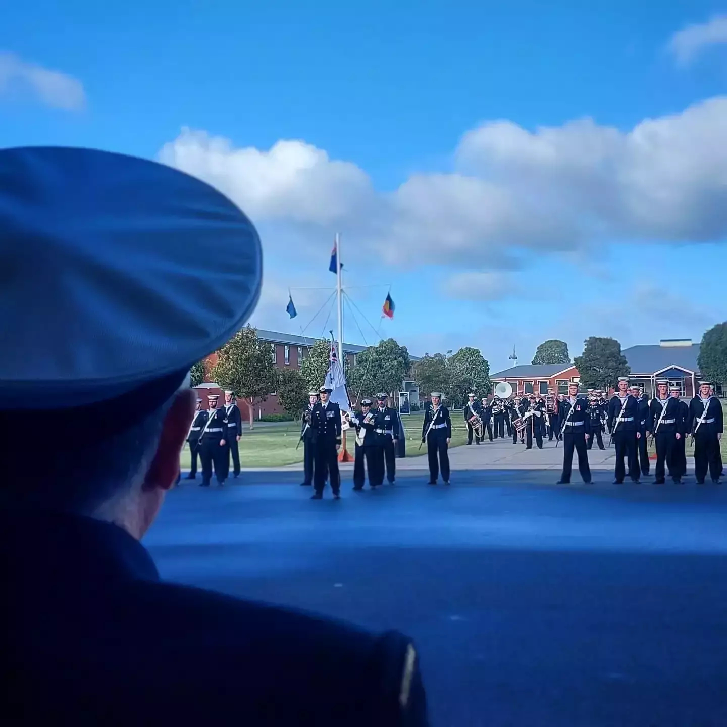 Auto-generated description: A group of uniformed individuals stand in formation on a parade ground, with flags and a person in a naval hat in the foreground.