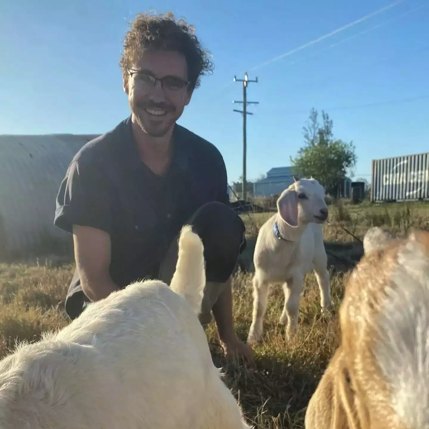 Auto-generated description: Joe is sitting on the grass petting goats on a farm with buildings and trees in the background.