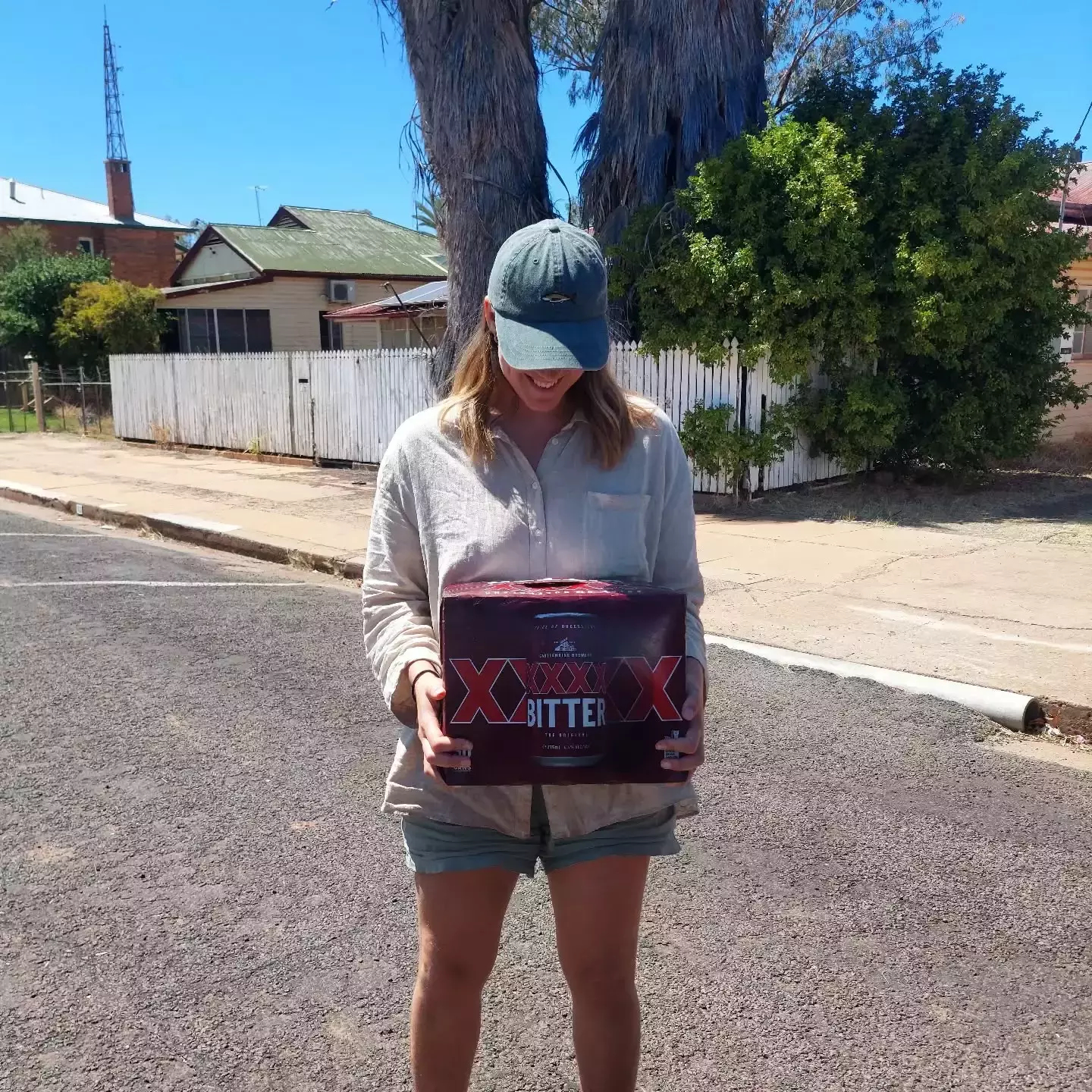 Auto-generated description: Tanja wearing a cap and holding a carton of XXXX Bitter stands on a street with houses and trees in the background.