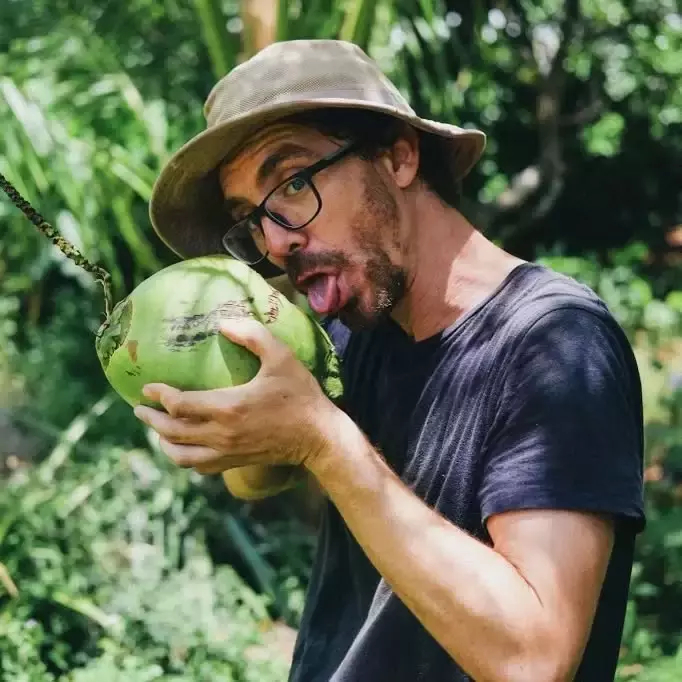 Auto-generated description: A man wearing glasses and a hat humorously pretends to lick a coconut in a lush outdoor setting.