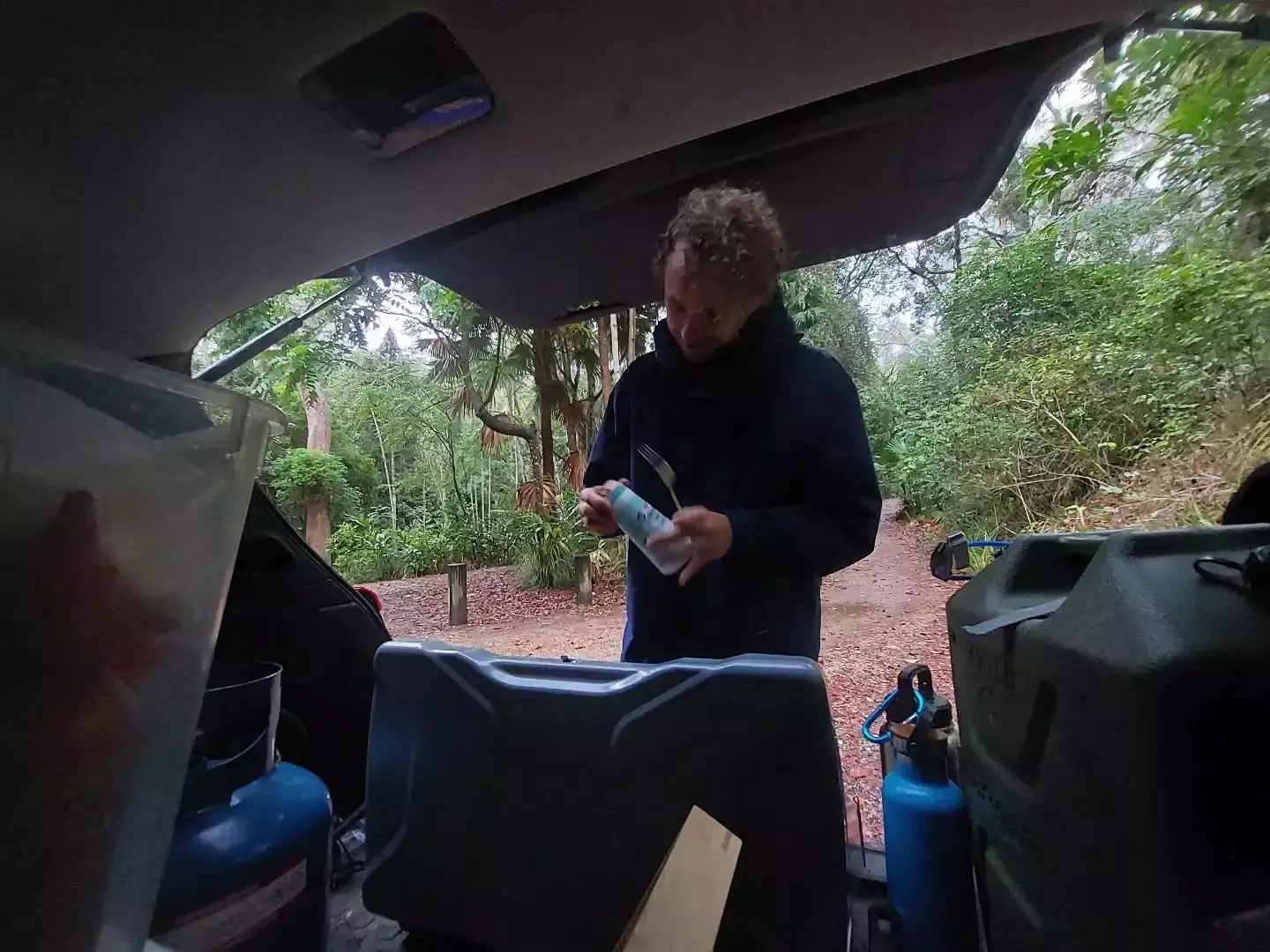 Auto-generated description: A person is cooking in the back of a vehicle, holding a container, with a forested area in the background.