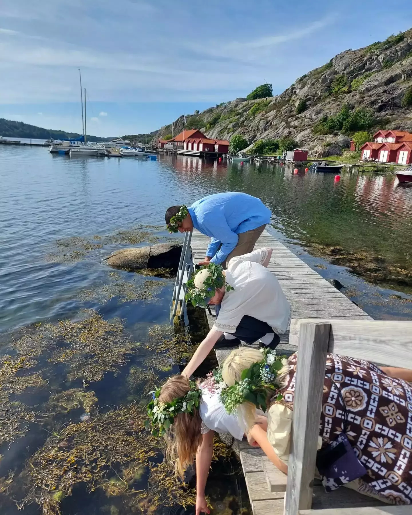 Auto-generated description: Four people, wearing floral wreaths, lean over a wooden dock to touch the water, with boats and rocky hills in the background.