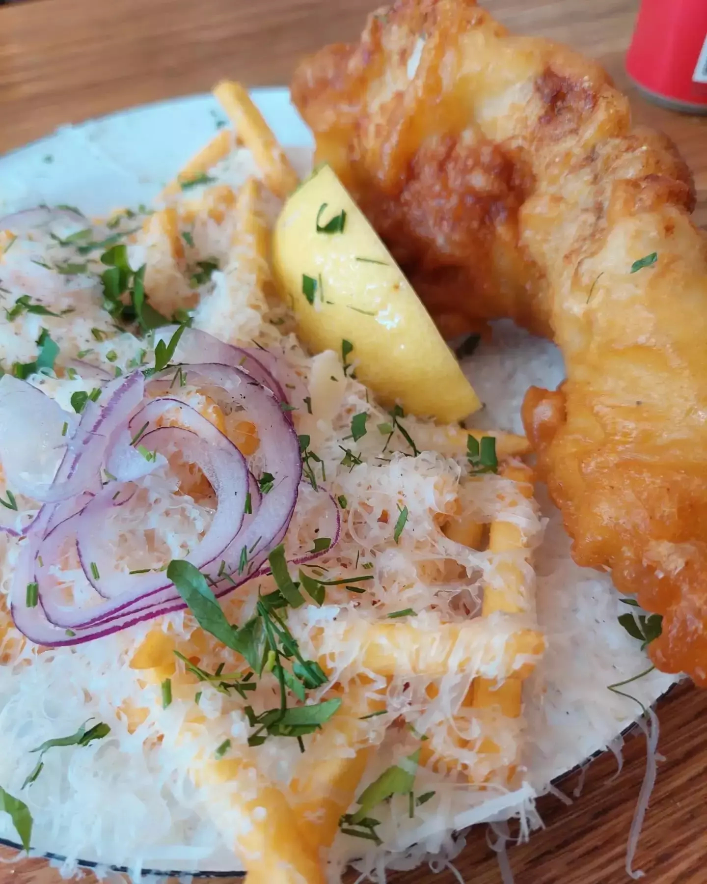 Auto-generated description: A plate features a crispy fish fillet with lemon, grated cheese, fresh herbs, red onion slices, and a pile of fries or potato sticks.