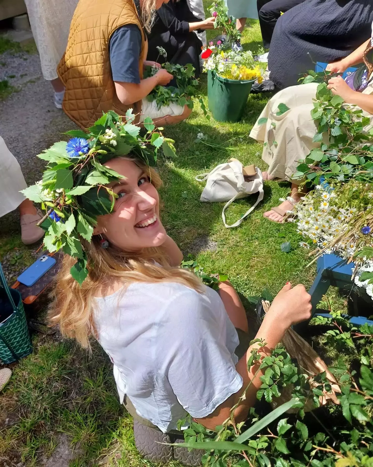 Auto-generated description: A smiling person is wearing a flower crown while sitting outdoors with others making floral arrangements.