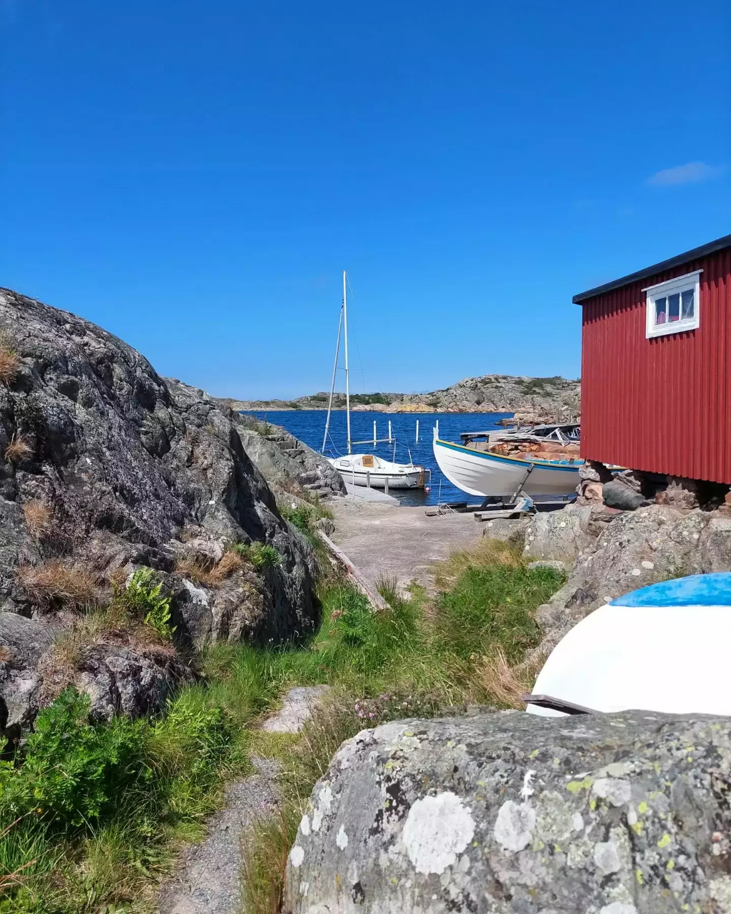 Auto-generated description: A small dock with boats is nestled between rocky terrain and a red wooden building, overlooking a calm sea under a clear blue sky.