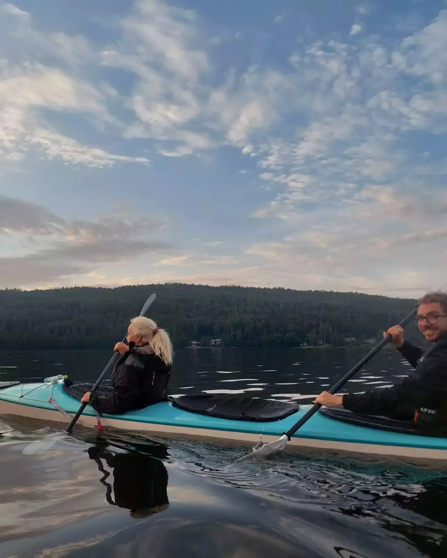 Auto-generated description: Joe and Annika are paddling a kayak on a calm lake surrounded by forested hills under a partly cloudy sky.