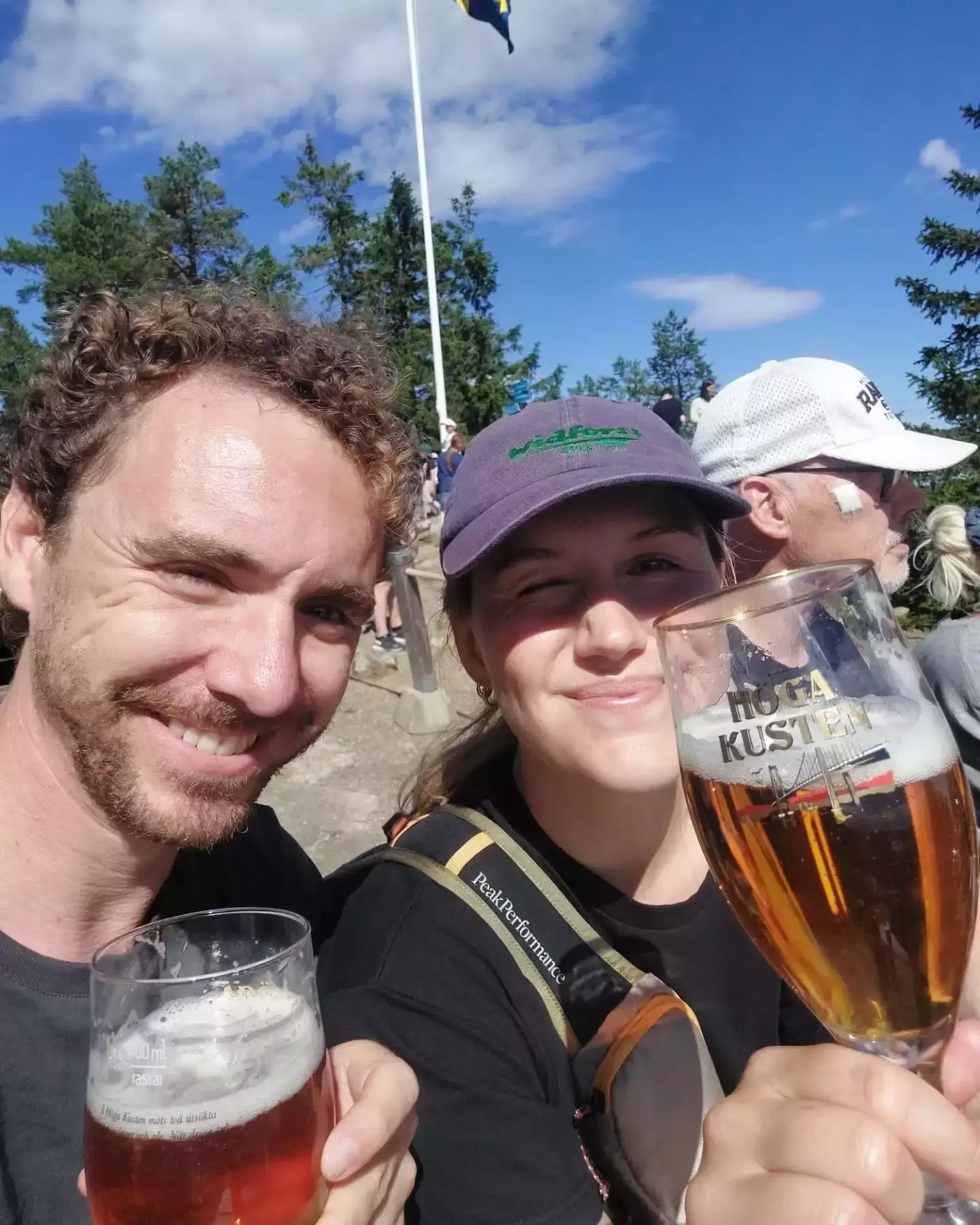 Auto-generated description: Tanja and Joe smiling and holding glasses of beer outdoors with a group of people and trees in the background.