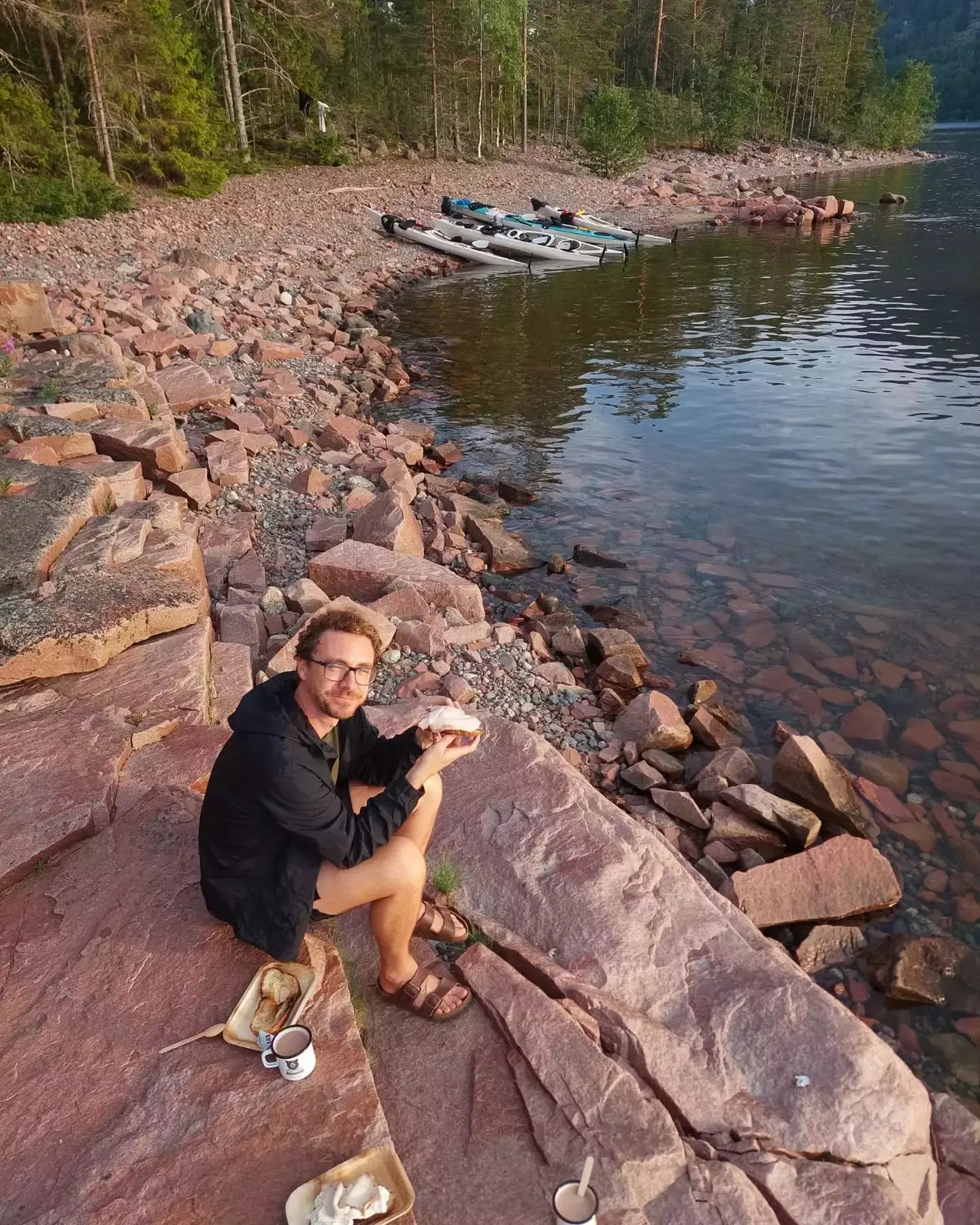 Auto-generated description: Joe sitting on a rocky shoreline enjoying a meal with kayaks and calm water in the background.