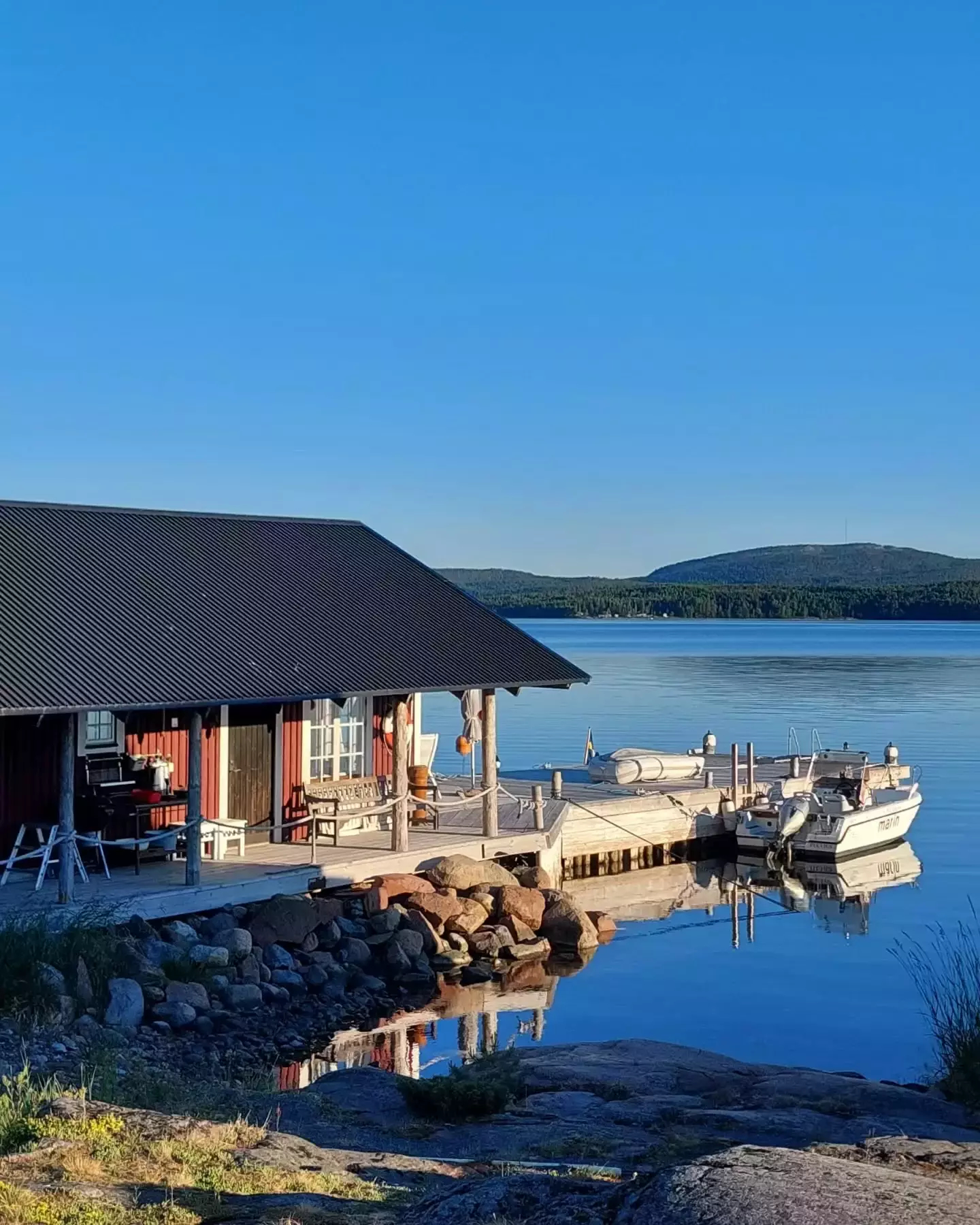 Auto-generated description: A serene lakeside scene features a wooden cabin, a small dock with boats, and a clear blue sky.