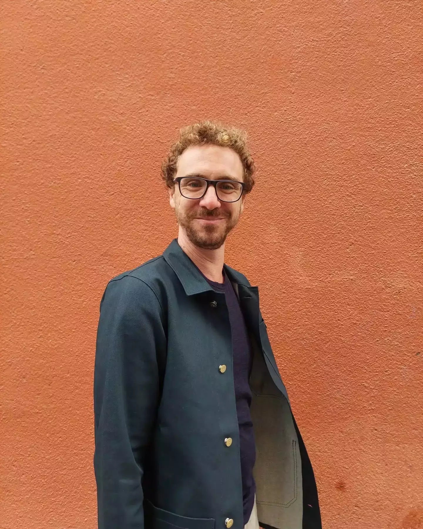 Auto-generated description: A person with curly hair, wearing glasses and a dark jacket, stands in front of a textured orange wall.