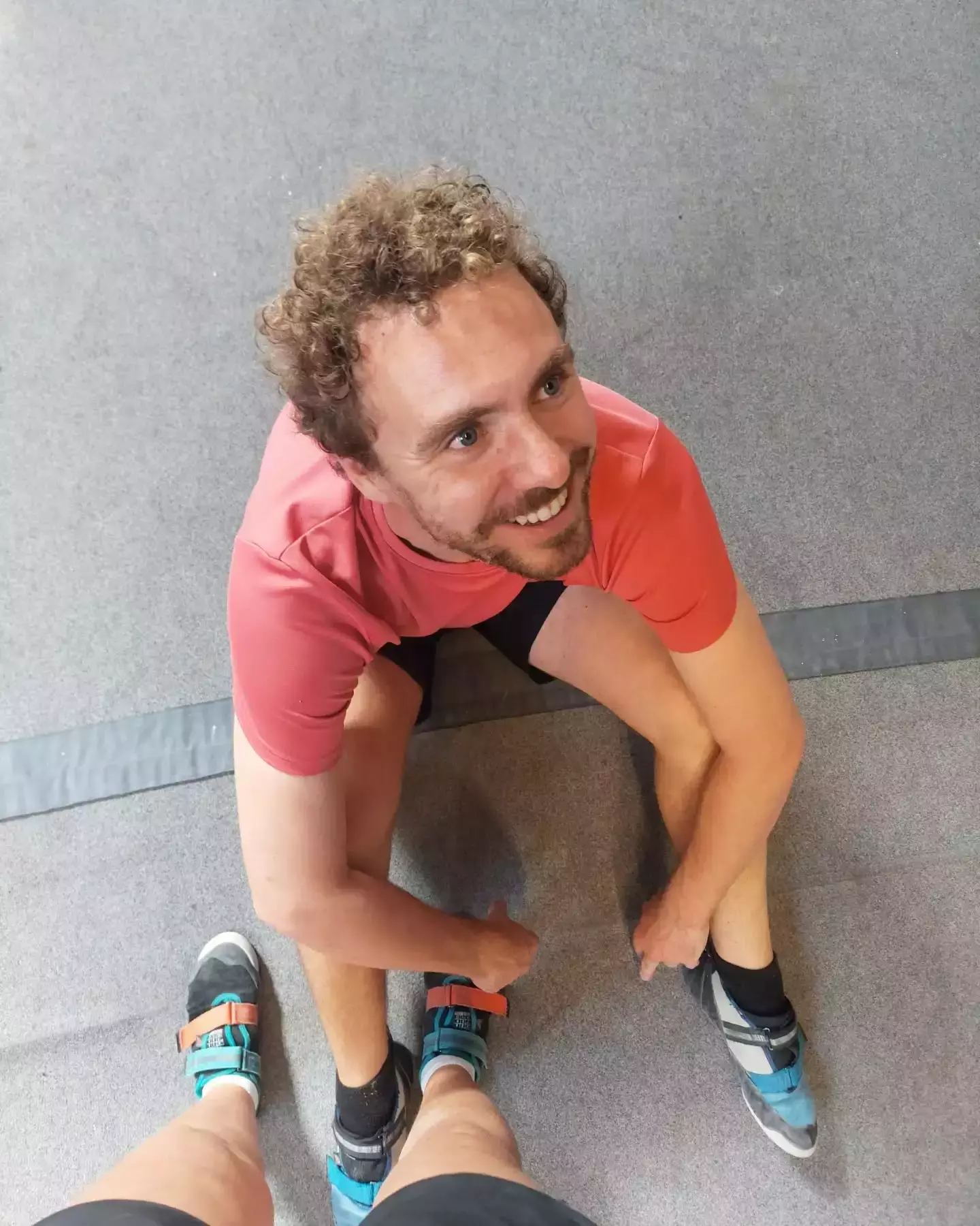 Auto-generated description: A person wearing a red shirt is sitting on the floor, looking up and smiling, with another person's feet visible in climbing shoes.