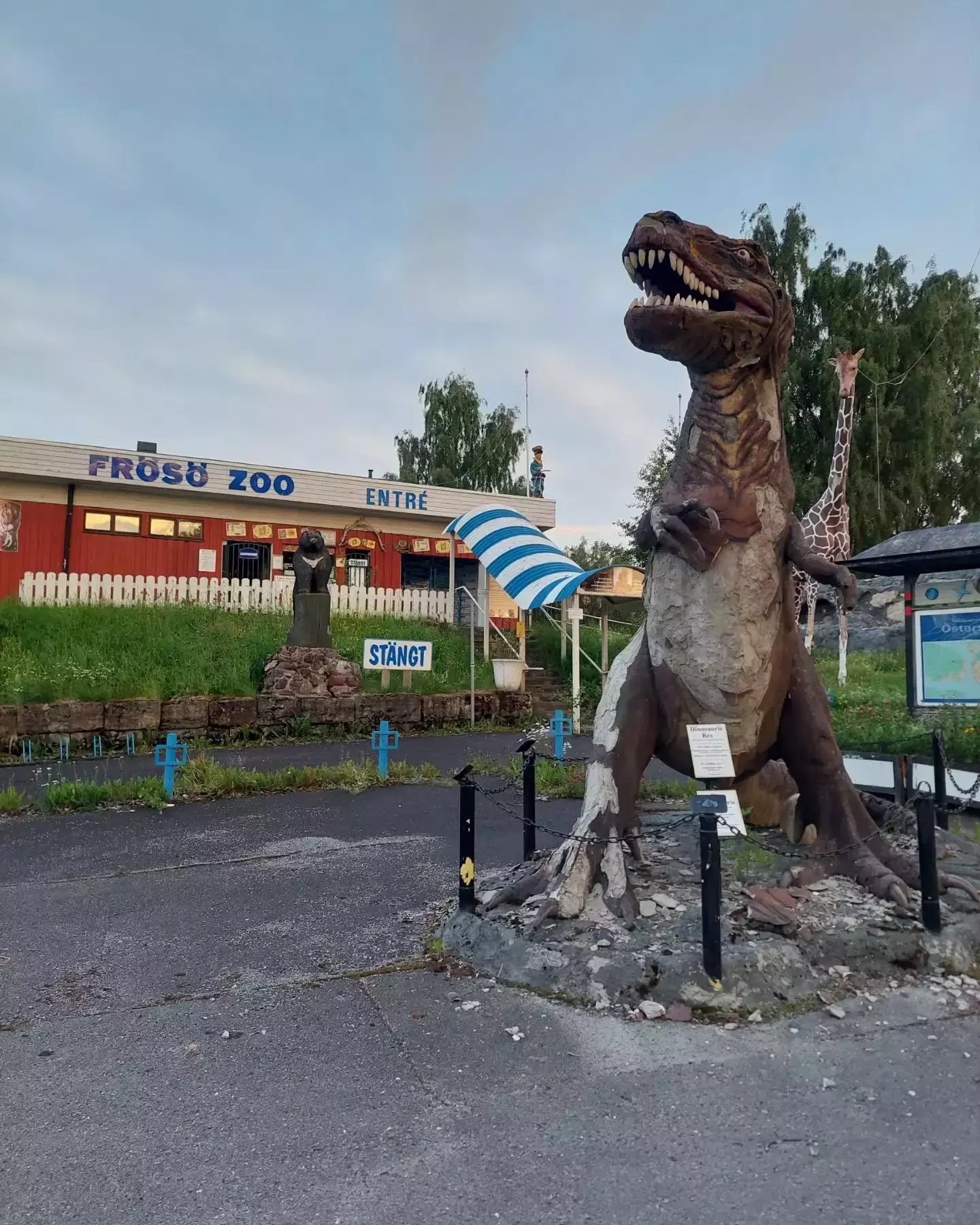 Auto-generated description: A dinosaur statue stands near the entrance of a zoo with a Stängt (closed) sign visible in the foreground.