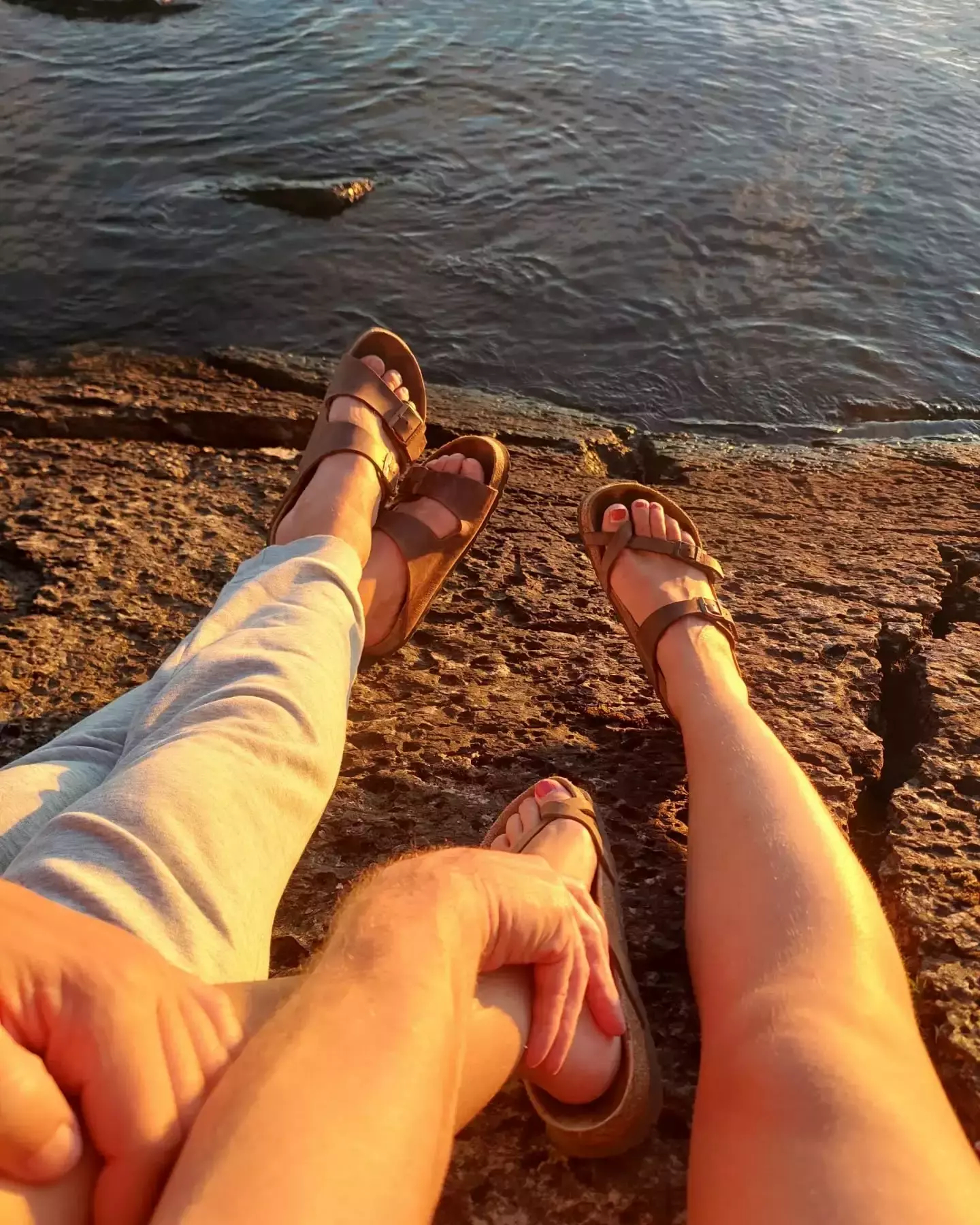 Auto-generated description: Two people are sitting on a rocky shore, with legs stretched out towards a body of water, enjoying a sunset.