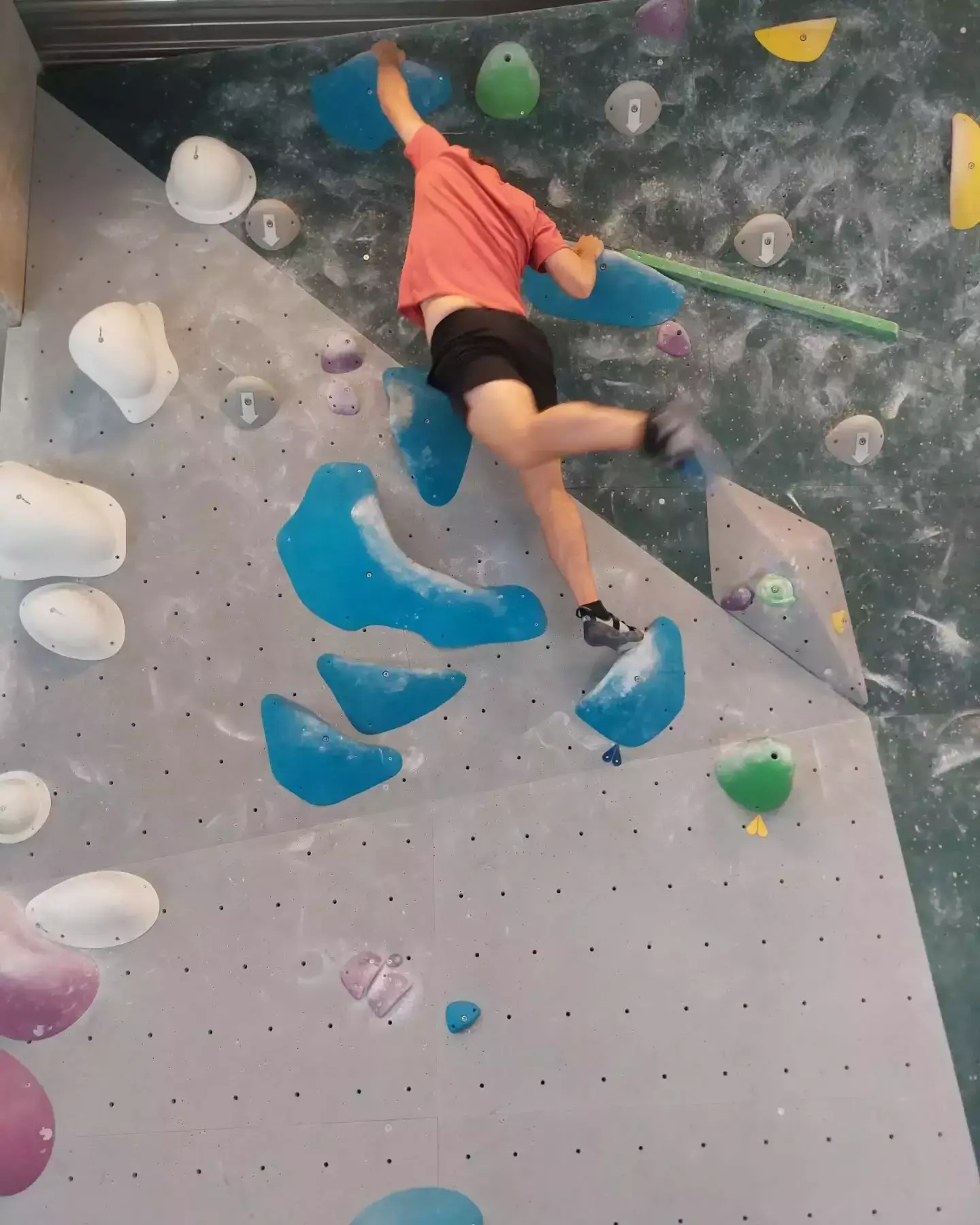 Auto-generated description: A person is climbing an indoor bouldering wall, reaching for a hold while wearing climbing shoes and a red shirt.