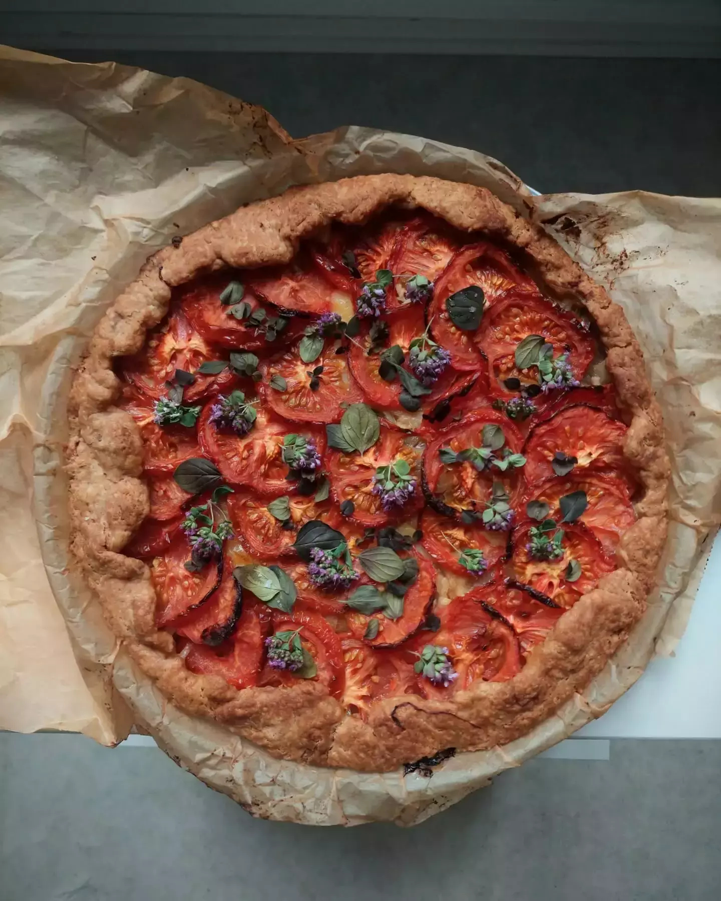 Auto-generated description: A savory tart featuring sliced tomatoes, fresh herbs, and a golden-brown crust is displayed on parchment paper.