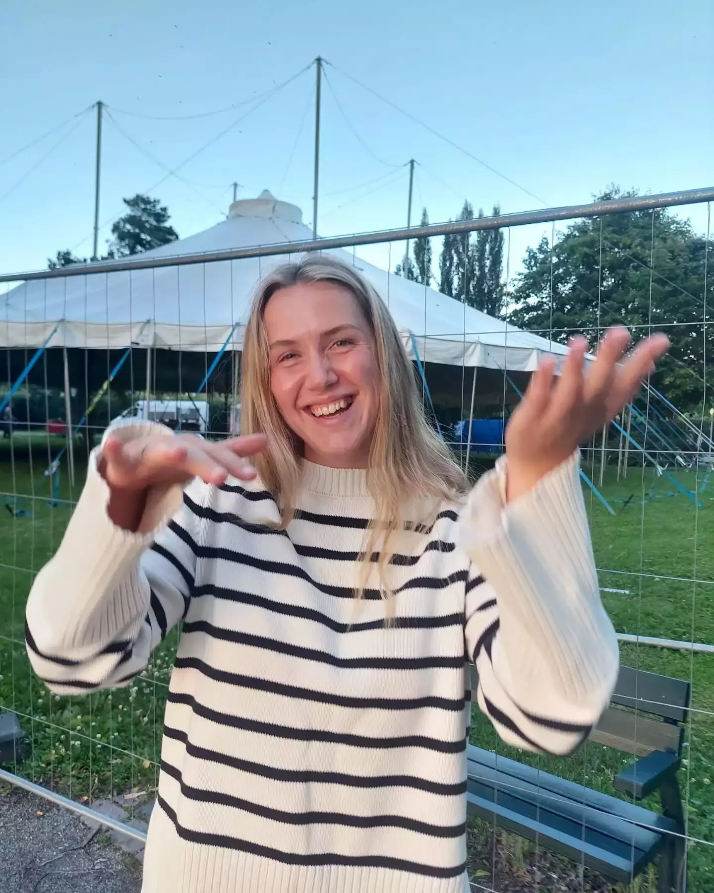 Auto-generated description: A person in a striped sweater is smiling and gesturing with their hands outdoors near a tent.