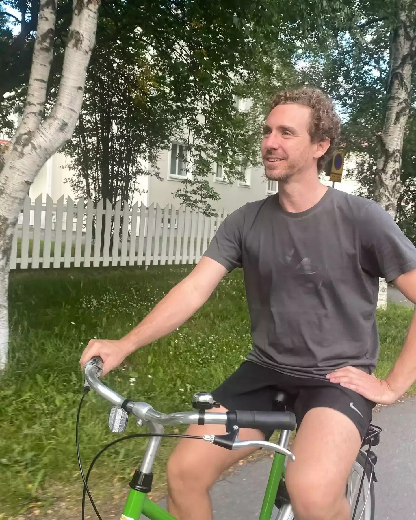 Auto-generated description: A person is riding a green bicycle along a path lined with trees and a white picket fence.