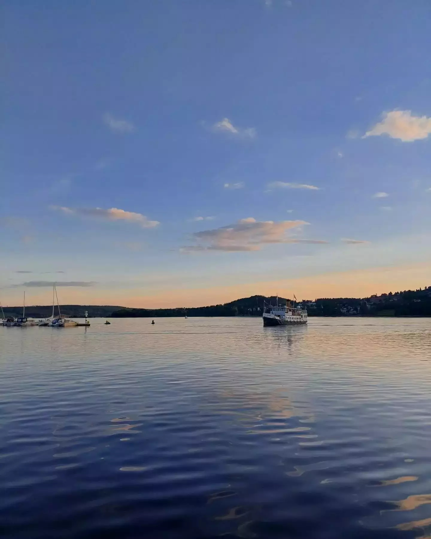 Auto-generated description: A serene lakeside scene features a few boats floating on calm water under a clear blue sky with scattered clouds and the distant silhouette of hills.