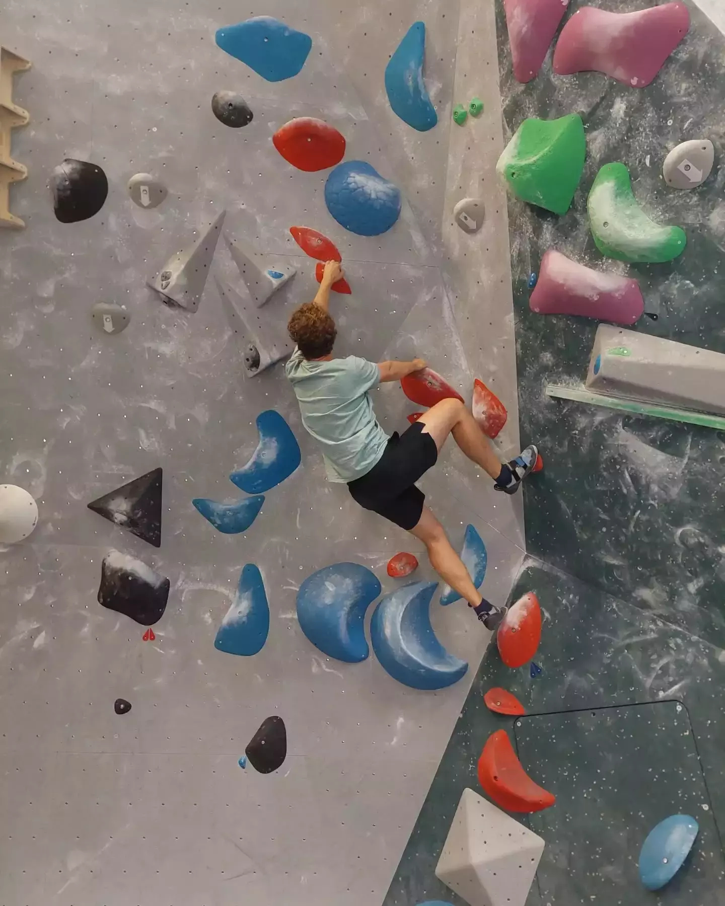 Auto-generated description: A person is bouldering indoors on a climbing wall with colorful handholds and footholds.