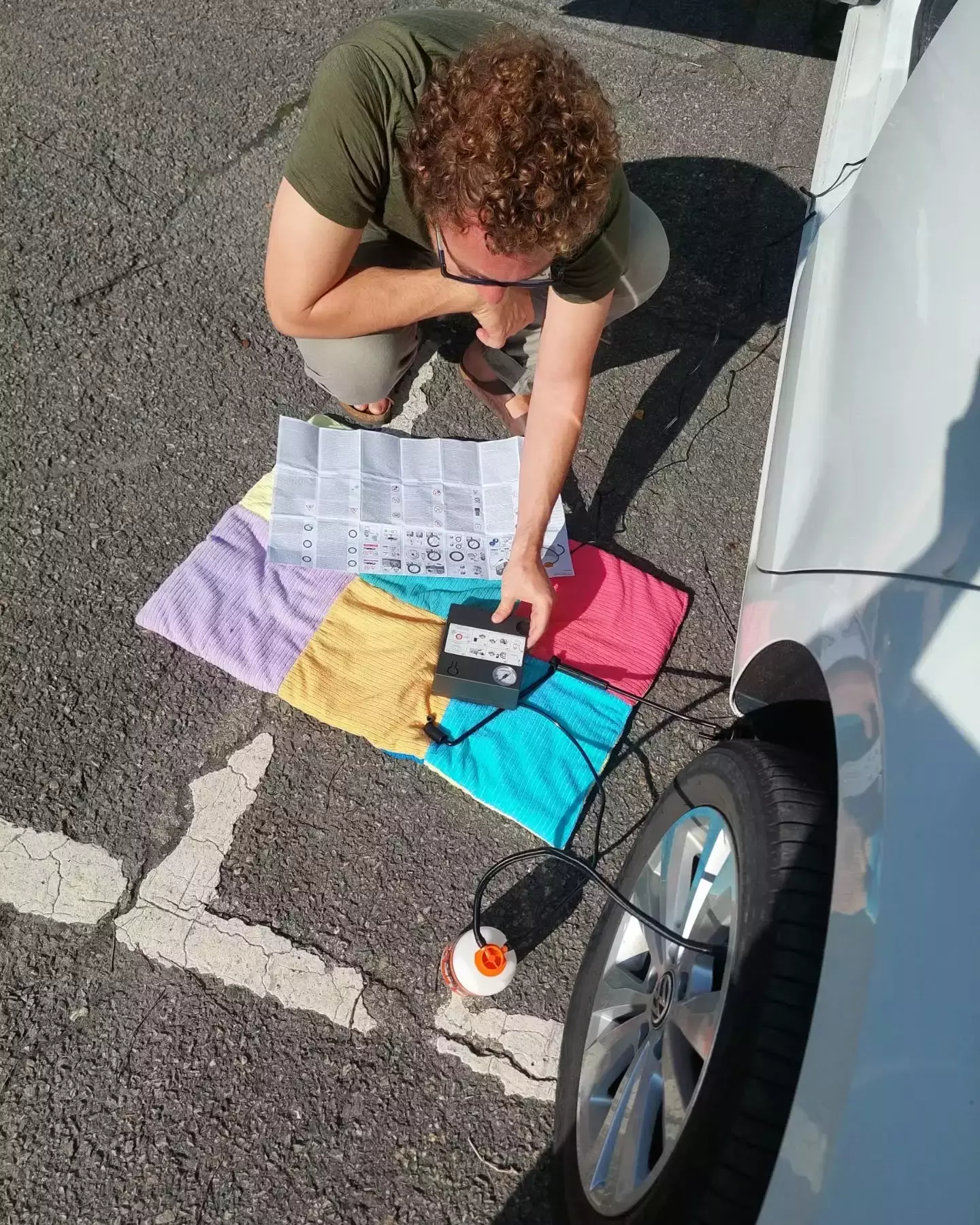 Auto-generated description: A person is kneeling on a colorful cloth while using a device to inflate a car tire in a parking lot.