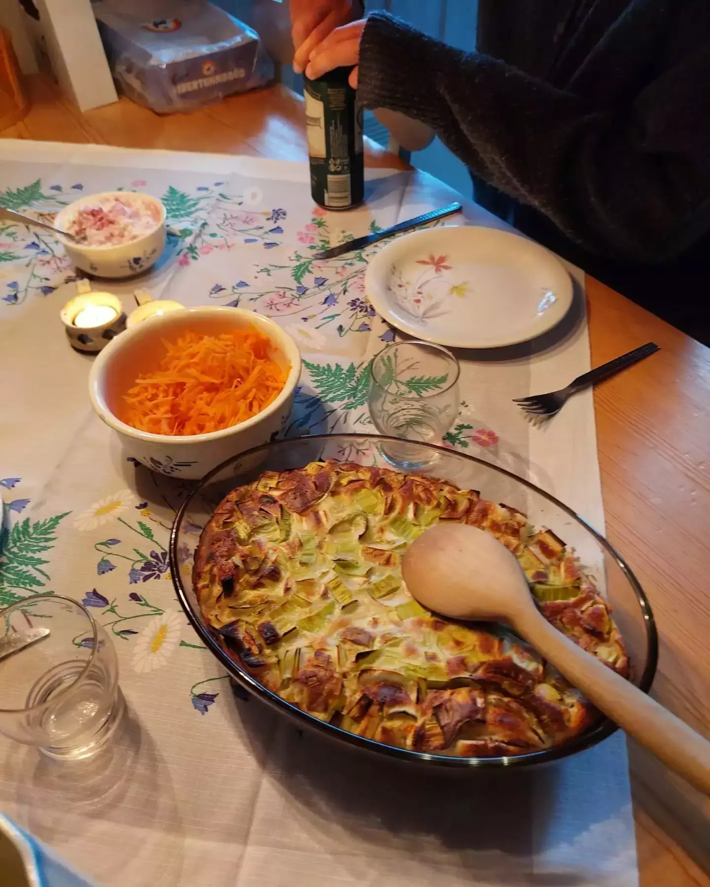 Auto-generated description: A table is set with a leek pie, a bowl of grated carrots, a plate, candles, and someone opening a bottle.