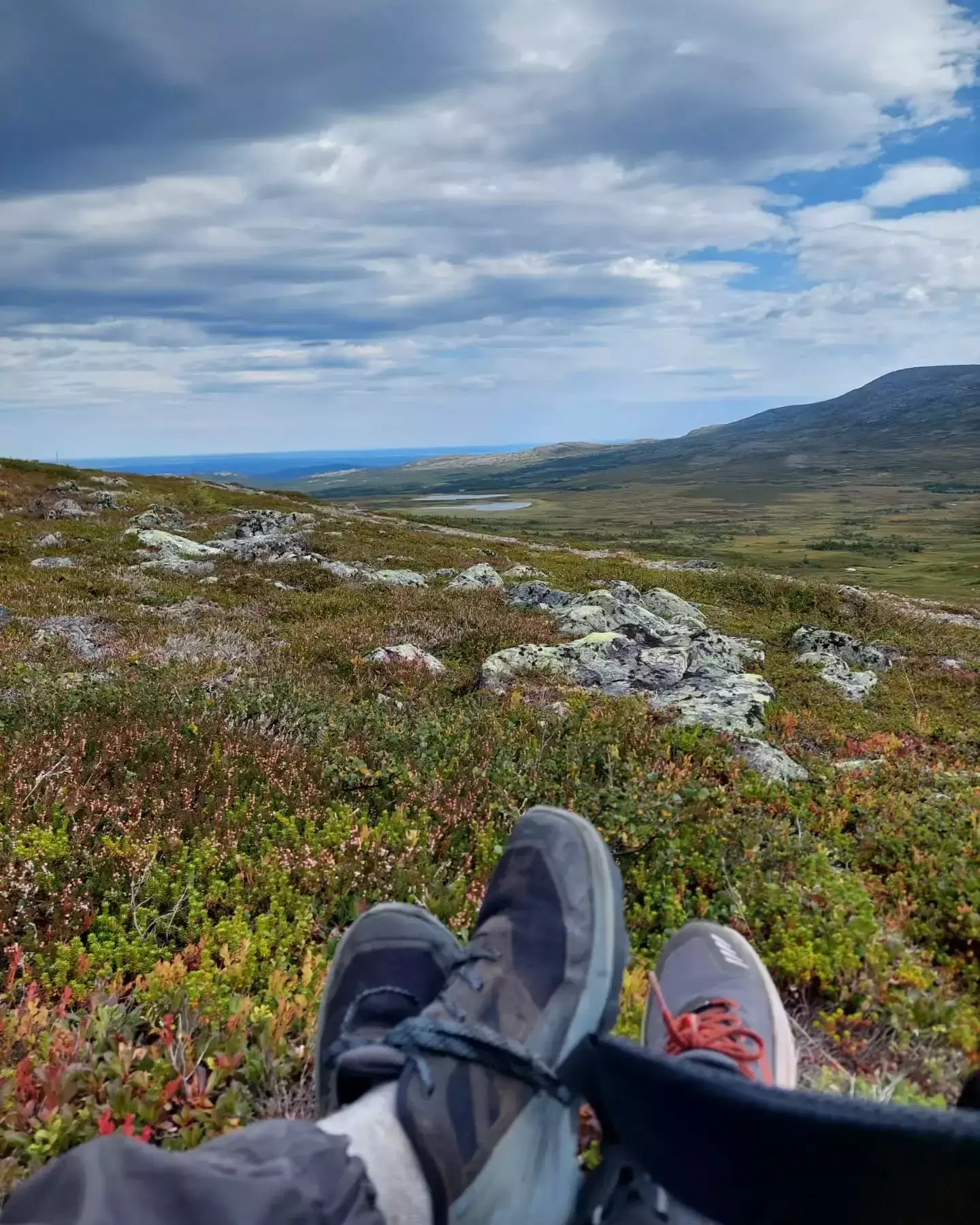 Auto-generated description: Two people are relaxing with their legs stretched out, overlooking a vast, scenic landscape with hills and cloudy skies.