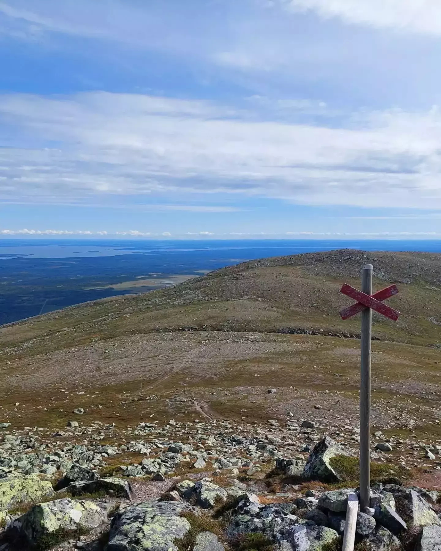 Auto-generated description: A rocky mountain landscape stretches towards a distant lake under a cloudy sky, with a wooden post marked by a red X in the foreground.