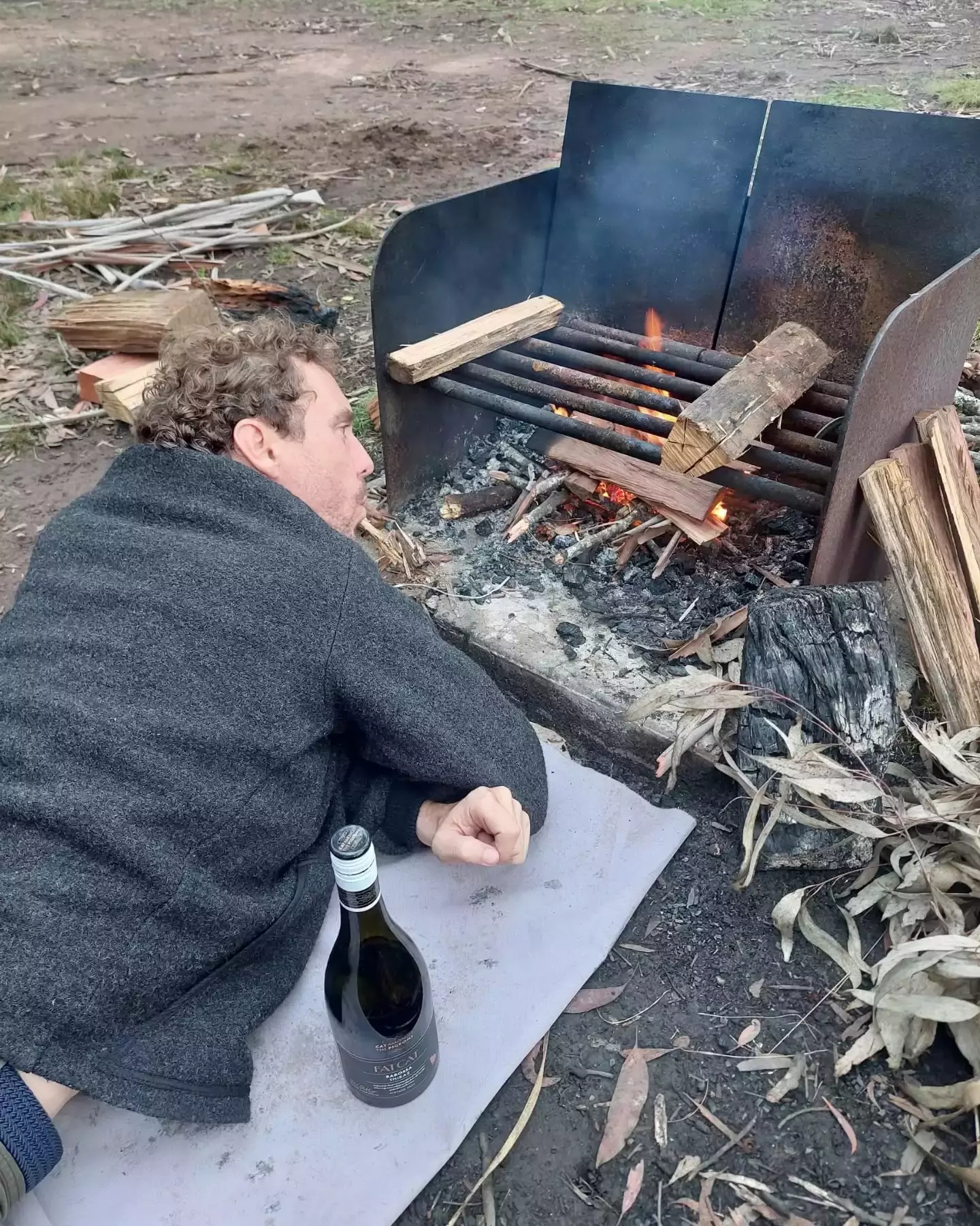 Joe lying on the ground blowing on a small fire.