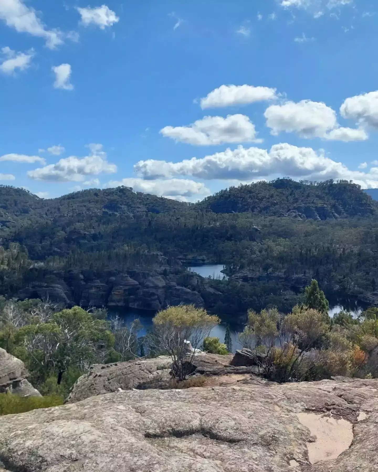 Auto-generated description: A scenic landscape features rocky terrain, dense green foliage, and a tranquil body of water under a partly cloudy sky.