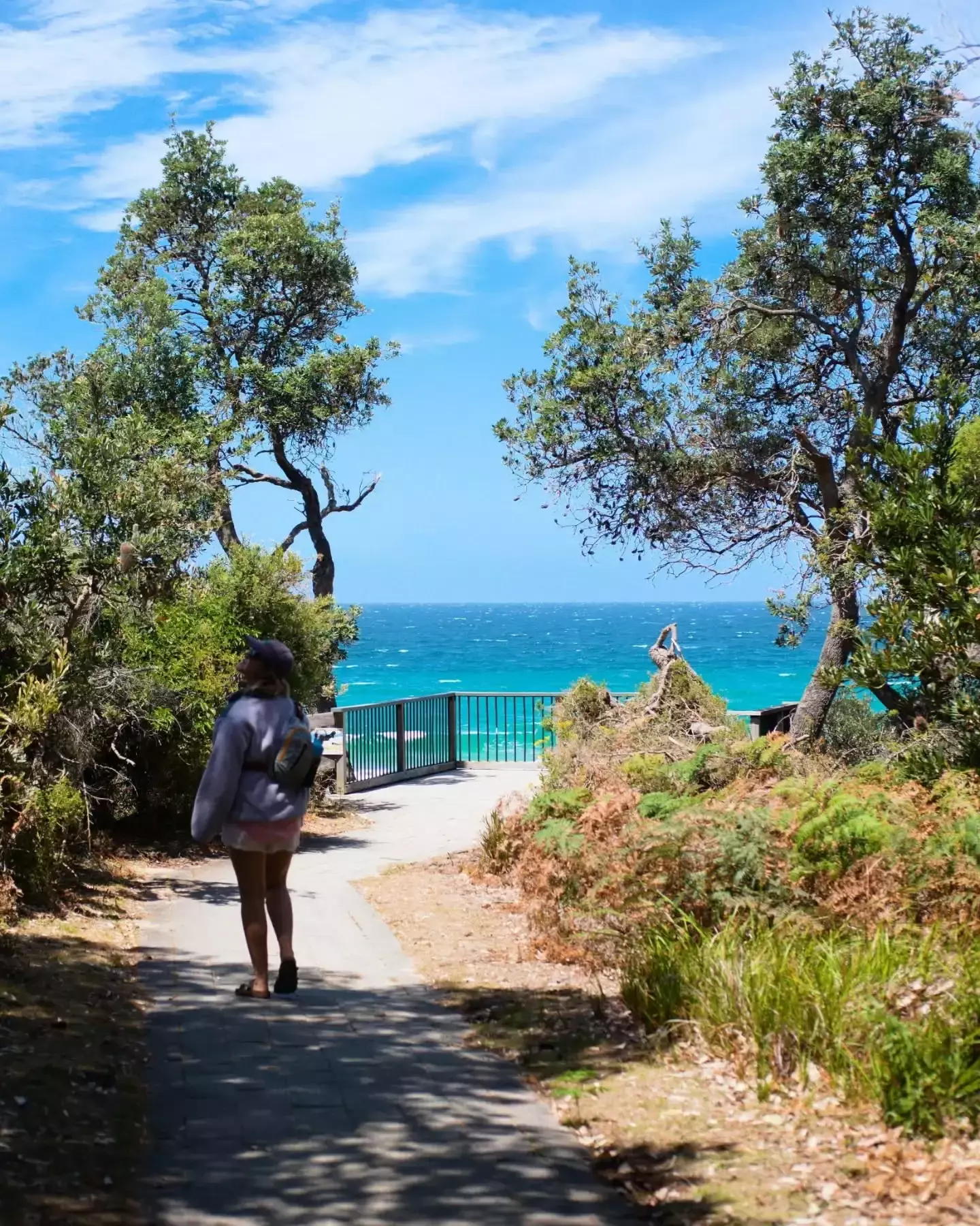 Auto-generated description: A person walks down a path surrounded by lush greenery, leading to a scenic ocean view under a partly cloudy sky.