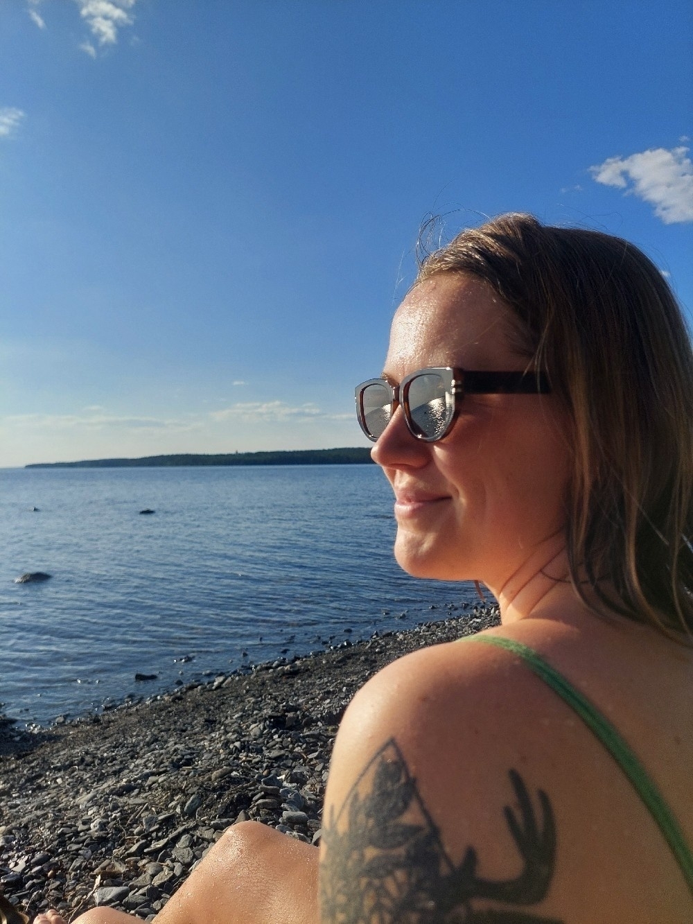 A person with sunglasses and a tattoo on their shoulder sits by the water on a sunny day.